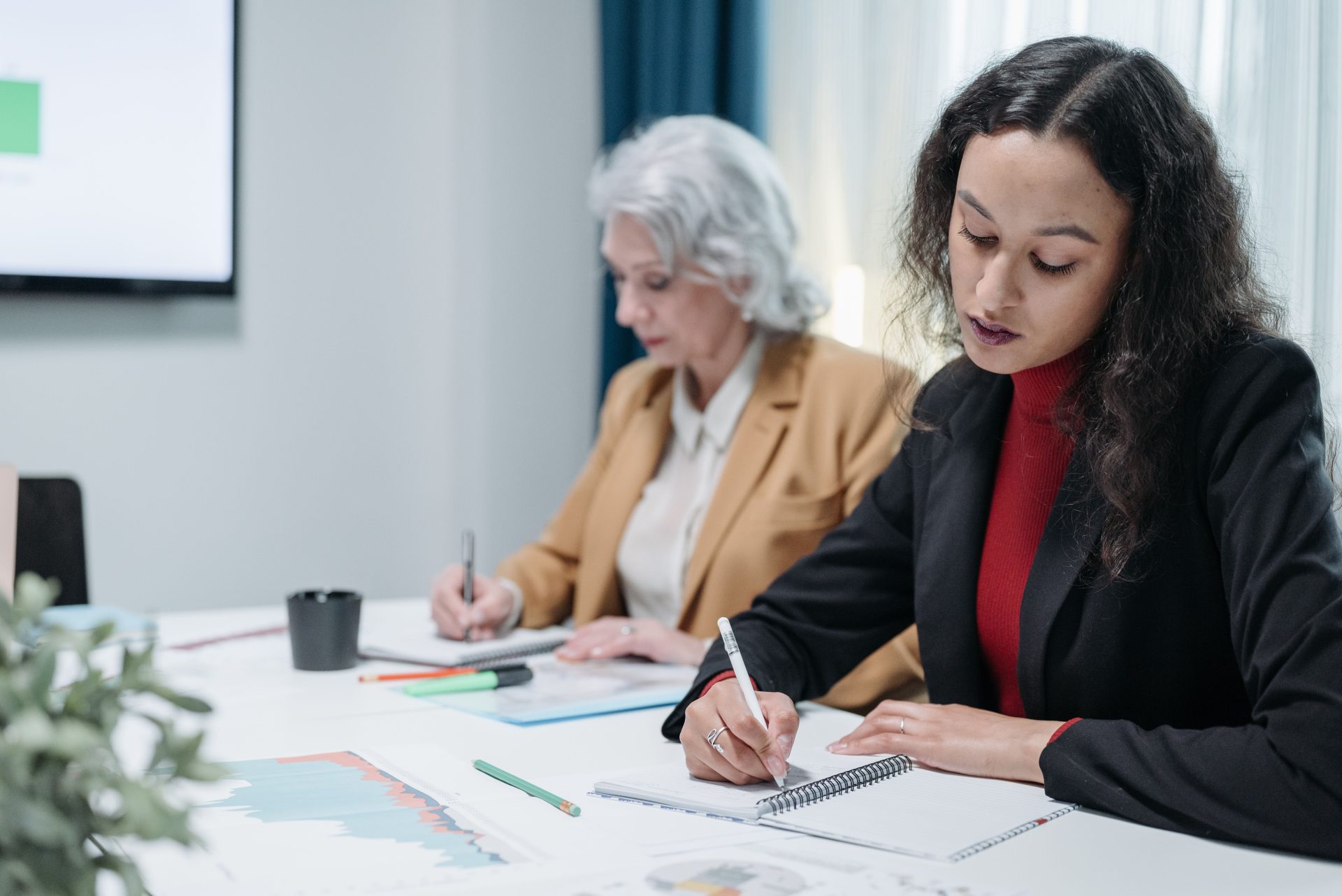 Two businesswomen engaged in work, taking notes during a meeting in a professional office environment.